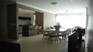 A spacious living area with a dining table and chairs, a kitchen area with a stove and cabinets, and a large window providing natural light. - Nice apartment, right on the beach and in a great location. (Meia Praia Itapema)