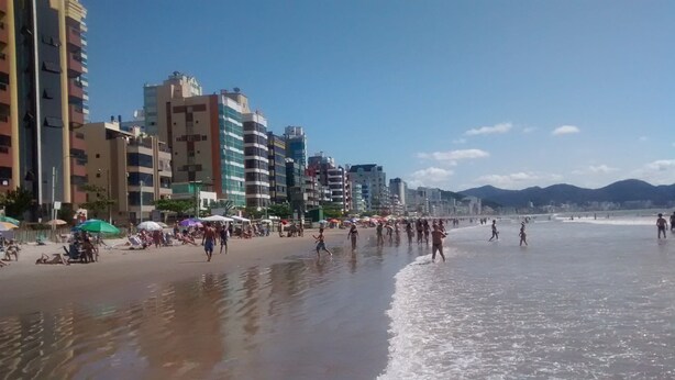 A beach with people walking and sunbathing, with buildings and mountains in the background. - Nice apartment, right on the beach and in a great location. (Meia Praia Itapema)