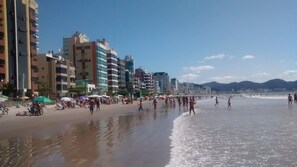 Una playa con gente caminando y tomando el sol, con edificios y montañas al fondo.