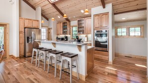 A spacious kitchen with wooden cabinets, a central island with bar stools, and a high ceiling with exposed beams. - Stoney Point by Seaside Vacations & Sales (Chincoteague Island)