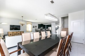 A modern dining area with a long table and chairs, a kitchen in the background, and a staircase leading to another level. - 1571 Observer Lane (Davenport)