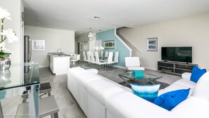 A modern living room with a white sofa, glass coffee table, and a TV. There is a dining area with white chairs and a kitchen in the background. - 1573 Plunker Drive (Davenport)