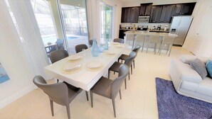 Un'elegante zona pranzo con un tavolo bianco apparecchiato per sei, circondato da sedie scure. Si intravede sullo sfondo una cucina con mobili scuri ed elettrodomestici in acciaio inossidabile.