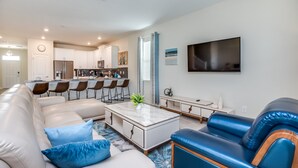 A modern living room with a sofa, a coffee table, and a blue leather chair. There is a kitchen in the background with a dining area. - 1670 Moonvalley Drive (Davenport)