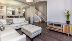 Un salon spacieux avec un canapé blanc, un grand pouf et un meuble de rangement gris. Un escalier avec une rampe blanche mène à l’étage supérieur.