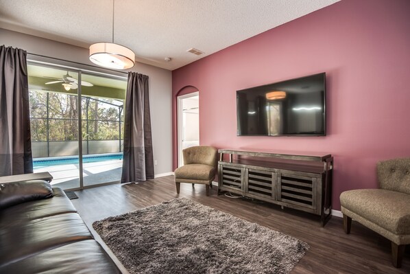 Una sala de estar moderna con un televisor grande, un sofá, una silla y vista a una piscina a través de un ventanal.