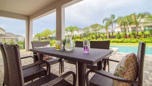 A covered patio area with wicker furniture, a table with drinks, and a view of a swimming pool and palm trees. - 281 Falls Drive (Kissimmee)