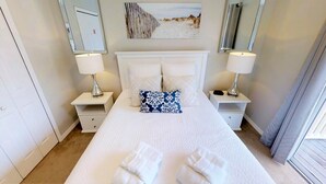 A neatly made bed with white linens and two pillows, one with a blue pattern. There are two bedside tables with lamps and a small drawer. A painting of a beach scene hangs above the bed. - 8523 Crystal Cove Loop (Kissimmee)