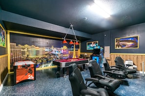 Mesa de bilhar em sala com mural da paisagem urbana, tacos de bilhar e cadeiras de jogos pretas.