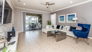 A modern living room with a white sofa, a blue armchair, a glass coffee table, and a ceiling fan. There is a dining area with a table and chairs visible through a glass door. - 2105 Morocco Way (Kissimmee)