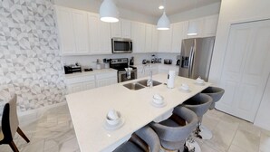 Una cocina moderna con armarios blancos, una isla central con taburetes, electrodomésticos de acero inoxidable e iluminación colgante.