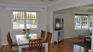 A dining area with a wooden table and chairs, a TV on a stand, and large windows with blinds. - Witton Bluff Cottage (christies beach)