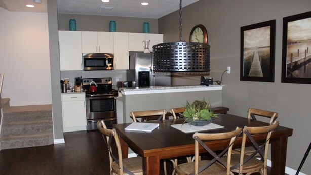 A dining area with a wooden table and chairs, a kitchen in the background, and a clock on the wall.