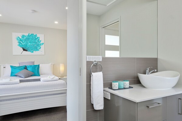 A modern hotel room with a bed, a large mirror, a white sink, and a teal artwork on the wall. - Beach View Apartment (BLACKMANS BAY)