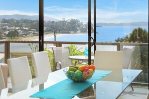 A dining area with a table, chairs, and a bowl of fruit. A view of the ocean and hills is visible through large windows. - Beach View Apartment (BLACKMANS BAY)