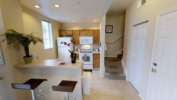 A compact kitchen with a refrigerator, microwave, stove, and wooden cabinets. There is a small island with two stools and a potted plant. A staircase leads to a door on the right. - Venetian Bay Villages Four-bedroom (Kissimmee)