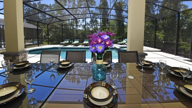 A dining table set for six with a vase of purple flowers, a glass table, and a view of a swimming pool and trees. - Watersong Resort Six-bedroom (Davenport)