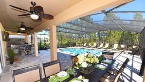 Un patio al aire libre con una mesa de comedor dispuesta para cuatro personas, rodeado de sillas, una piscina y un área de estar techada con sillones.