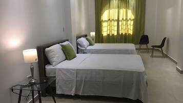 Two single beds with white bedding and green pillows, each with a bedside table and lamp, are positioned in a clean and minimalist space with a large window and air conditioning unit.
