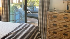 Un dormitorio con cama, cómoda de mimbre, lámpara y vistas a un patio exterior con sillas y mesa.