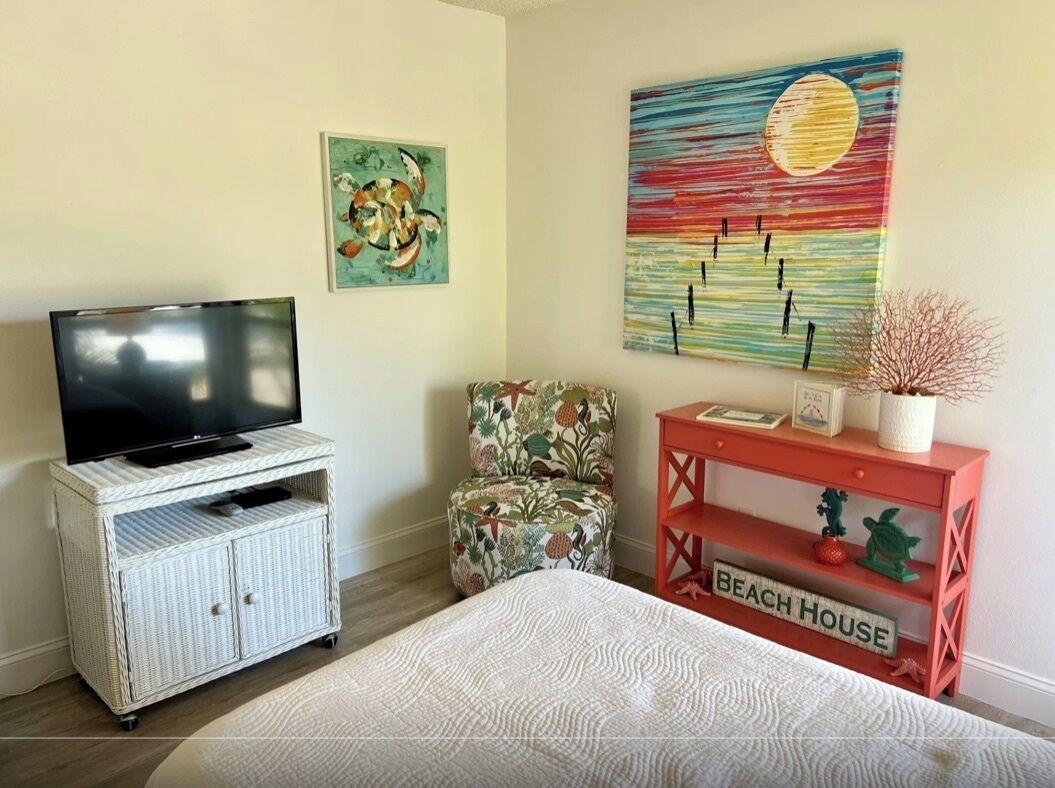Une chambre avec un lit, un téléviseur posé sur un meuble blanc, un tableau coloré, une chaise et une petite table rouge sur laquelle est posé un panneau indiquant « MAISON DE PLAGE ».