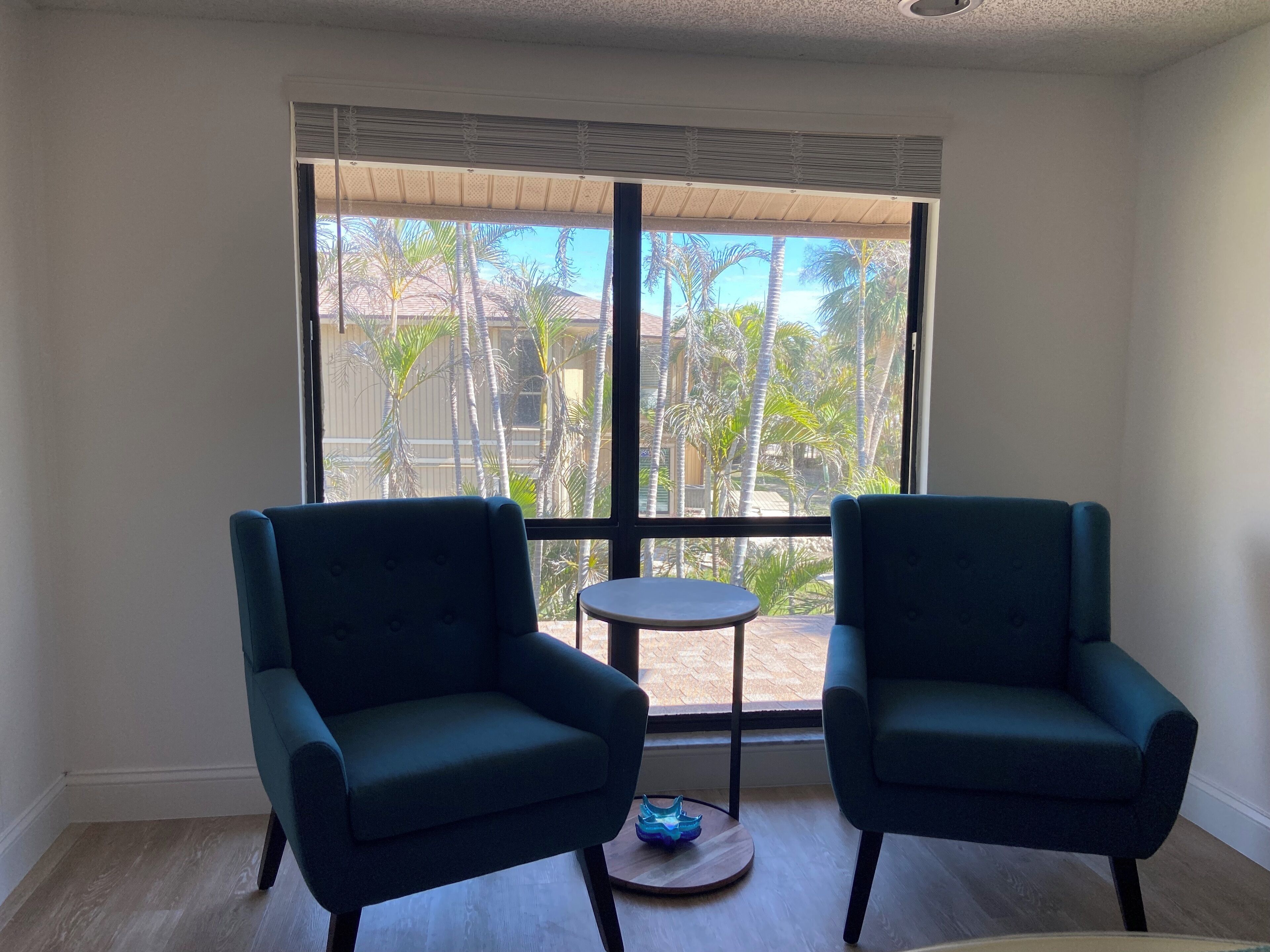 Una habitación con dos sillones azul oscuro, una mesita redonda y un ventanal con vistas a palmeras y un edificio.