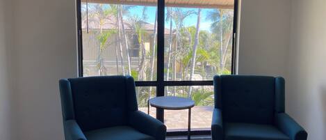 Una habitación con dos sillones azul oscuro, una mesita redonda y un ventanal con vistas a palmeras y un edificio.