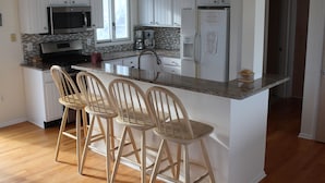Cocina con alacenas blancas, encimera de granito, microondas de acero inoxidable, refrigerador y tres taburetes de madera para barra.
