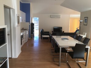 A modern kitchen with white cabinets and a built-in oven, a dining area with a white table and black chairs, and a living area with a black sofa and a TV. - Golf and Beach Retreat (Collingwood Park)
