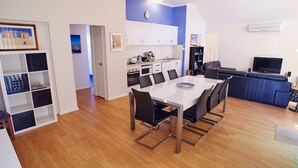 A modern kitchen with white cabinets, a dining area with a white table and black chairs, and a living area with a blue sofa and a flat-screen TV. - Golf and Beach Retreat (Collingwood Park)