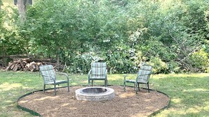 Een ronde vuurplaats omringd door drie stoelen met gestreepte kussens, gelegen in een tuin met een stenen rand en weelderige begroeiing op de achtergrond.