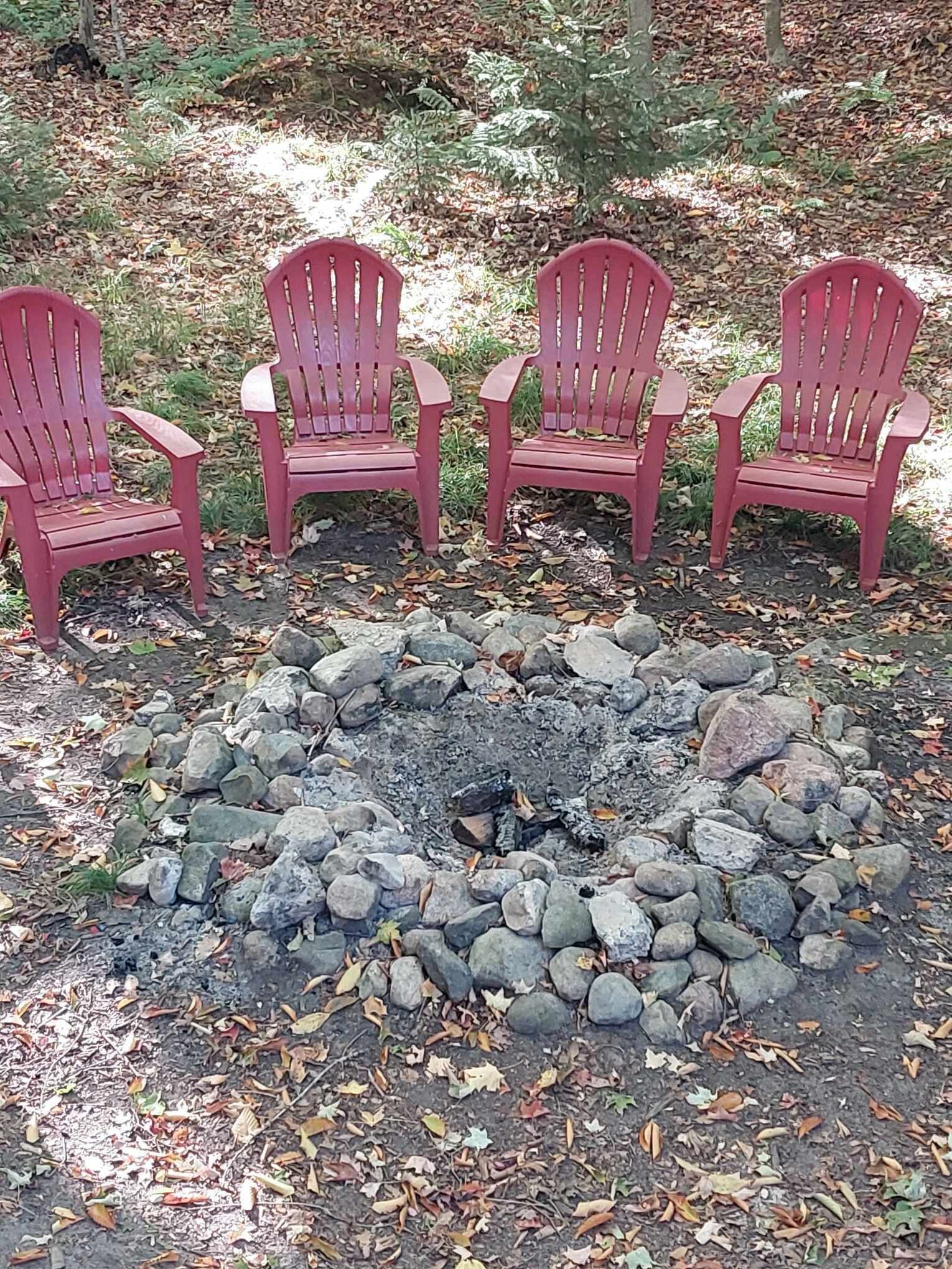 Cuatro sillas Adirondack rojas dispuestas alrededor de un hogar de piedra en un área boscosa.
