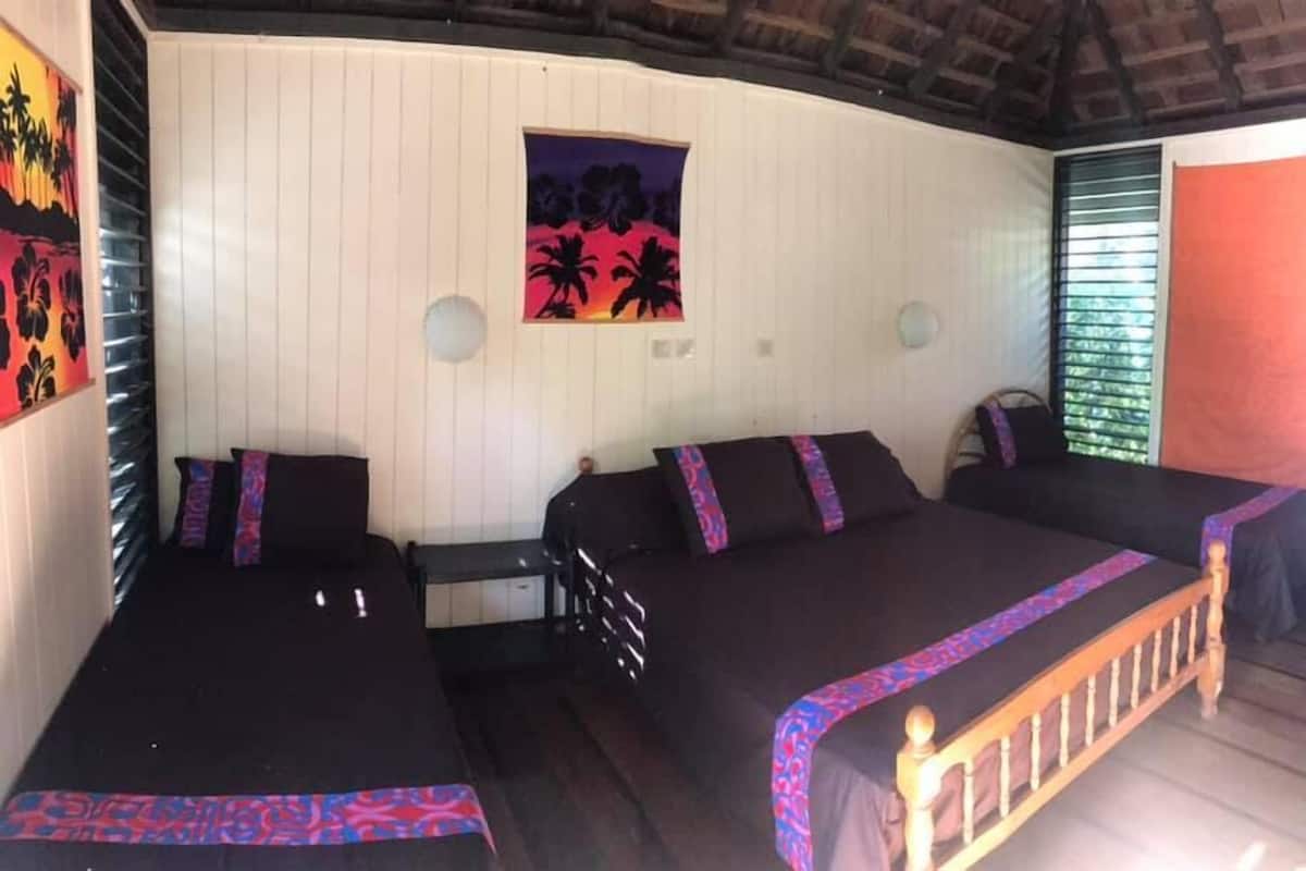A room with two beds, a wooden headboard, a wall with a tropical sunset painting, and a window with blinds.