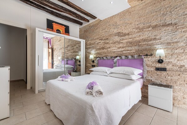 A bedroom with a large mirror, a bed with white and purple bedding, and a stone wall.