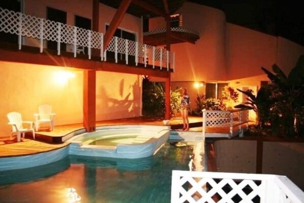 A swimming pool with a wooden deck, lounge chairs, and a covered area with a railing.