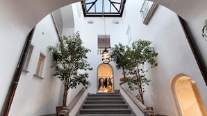 Un escalier surmonté d’une lucarne, bordé d’arbres et menant à un espace intérieur lumineux.