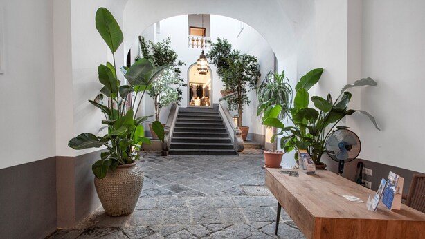 Un couloir dallé de pierre, avec une table en bois, des plantes en pot et un escalier menant à une pièce ornée d’un lustre.