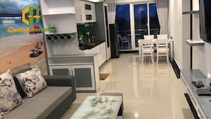 Una sala moderna con un sofá, una mesa de centro y una zona de comedor con sillas. Hay una cocina con armarios y un refrigerador. La habitación tiene un ventanal con cortinas.