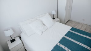 A neatly made bed with white bedding and a blue striped throw blanket, a white nightstand with a lamp, and a white wardrobe in a minimalist room. - Semi-detached house 5 'walk from the beach (La Garita, Telde, Las Palmas)