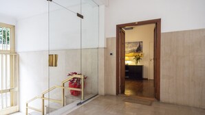 A modern hotel lobby with a glass elevator, a wooden staircase, and a reception area with a lamp and vase.