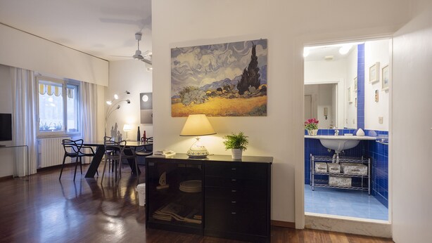 A modern living room with a dining area, a black cabinet with drawers, a painting on the wall, and a bathroom visible through an open door.