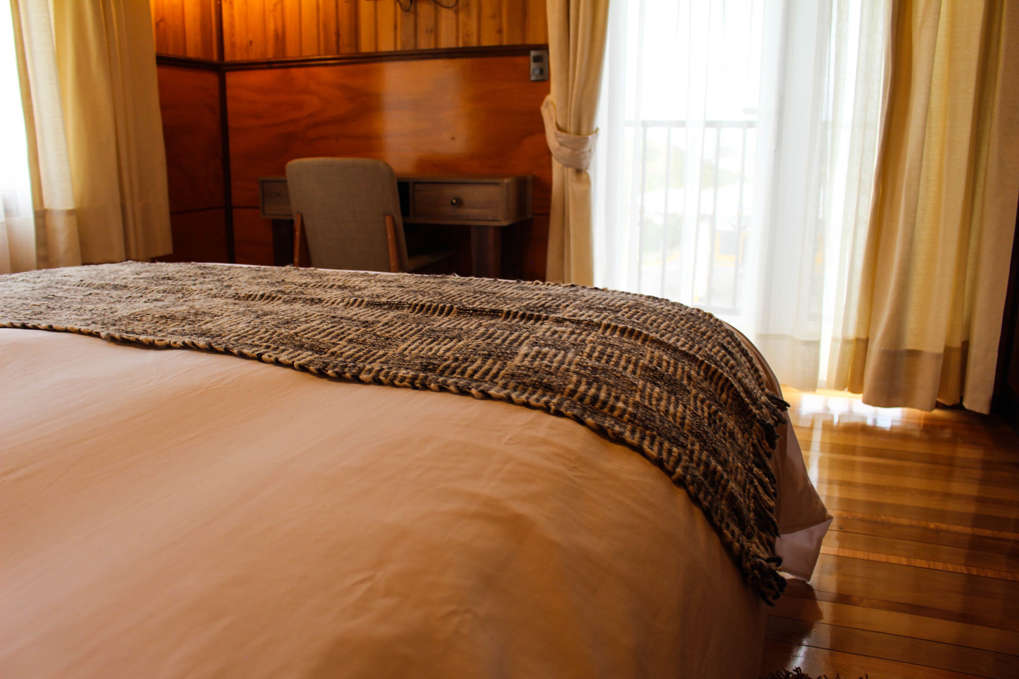 A neatly made bed with a patterned blanket, a wooden desk, and a chair in a room with wooden walls and large windows.