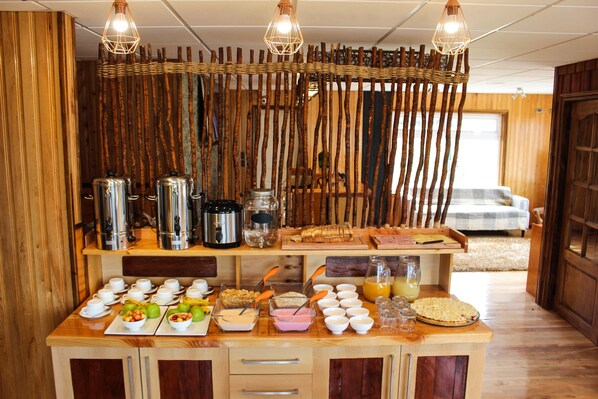 Un bufé de desayuno con diversos alimentos y bebidas, una estación de café y un separador de mimbre en una habitación con paredes de madera y una cama al fondo.