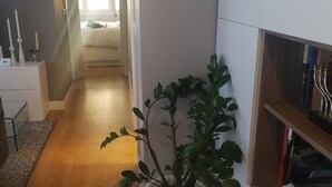Ein Flur mit Holzboden, einer weißen Topfpflanze, einem weißen Schrank mit Büchern und einem Bett im Hintergrund.