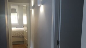 Ein schmaler Korridor mit Holzboden, einer Tür rechts und einem Bett im Hintergrund.