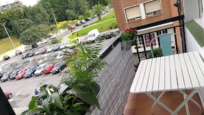 Ein Balkon mit weißem Tisch und Stühlen, Blumentöpfen und Blick auf einen Parkplatz mit verschiedenen Autos.