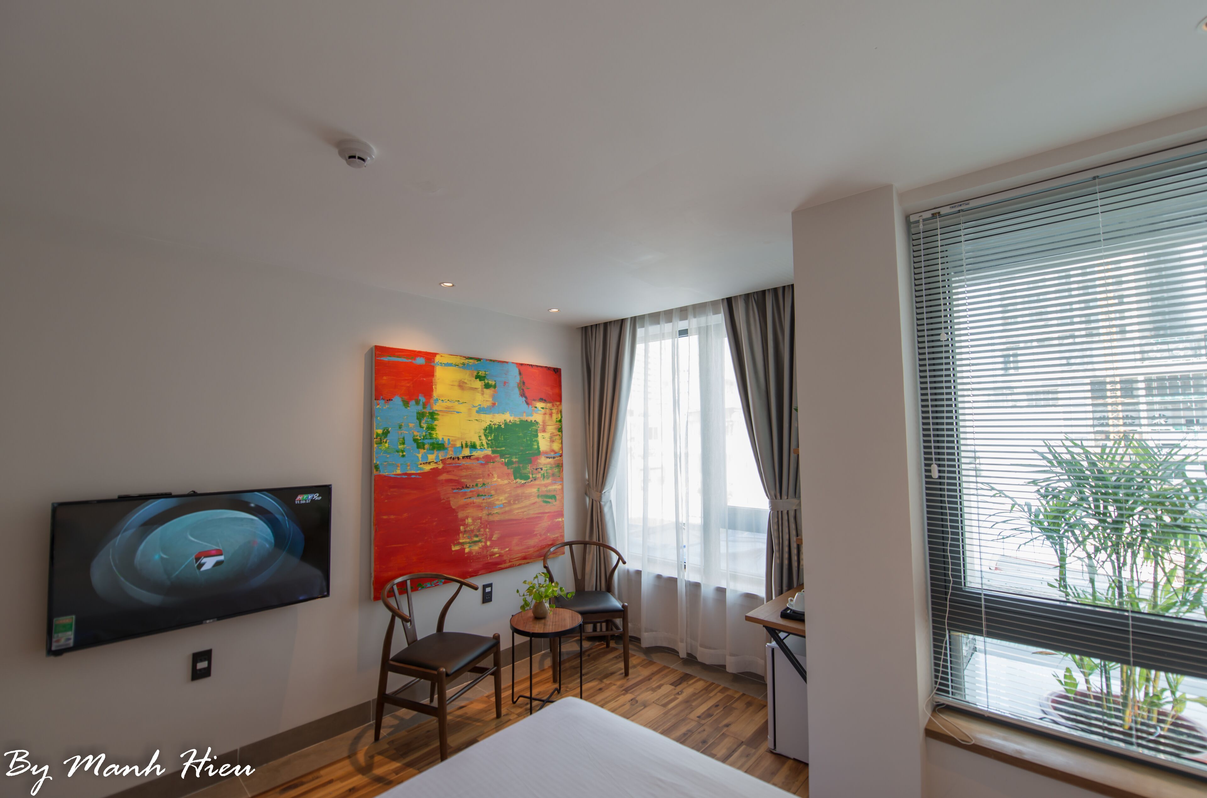 A hotel room with a bed, a TV mounted on the wall, a desk with a chair, a small table with a plant, and a window with blinds.