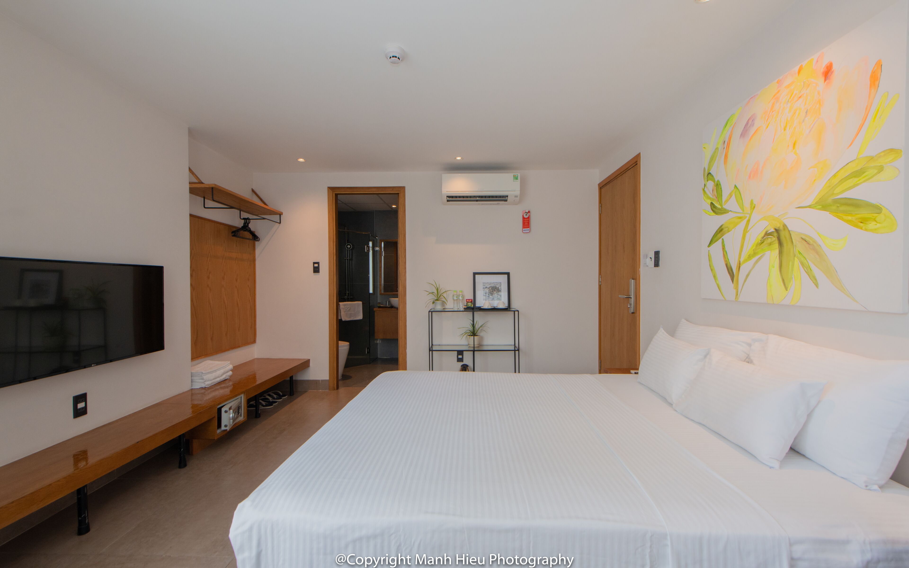 A hotel room with a large bed, a TV mounted on the wall, a wooden bench, and a plant on a small table.