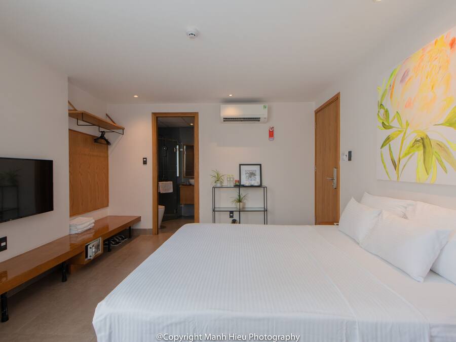 A hotel room with a large bed, a TV mounted on the wall, a wooden bench, and a plant on a small table.