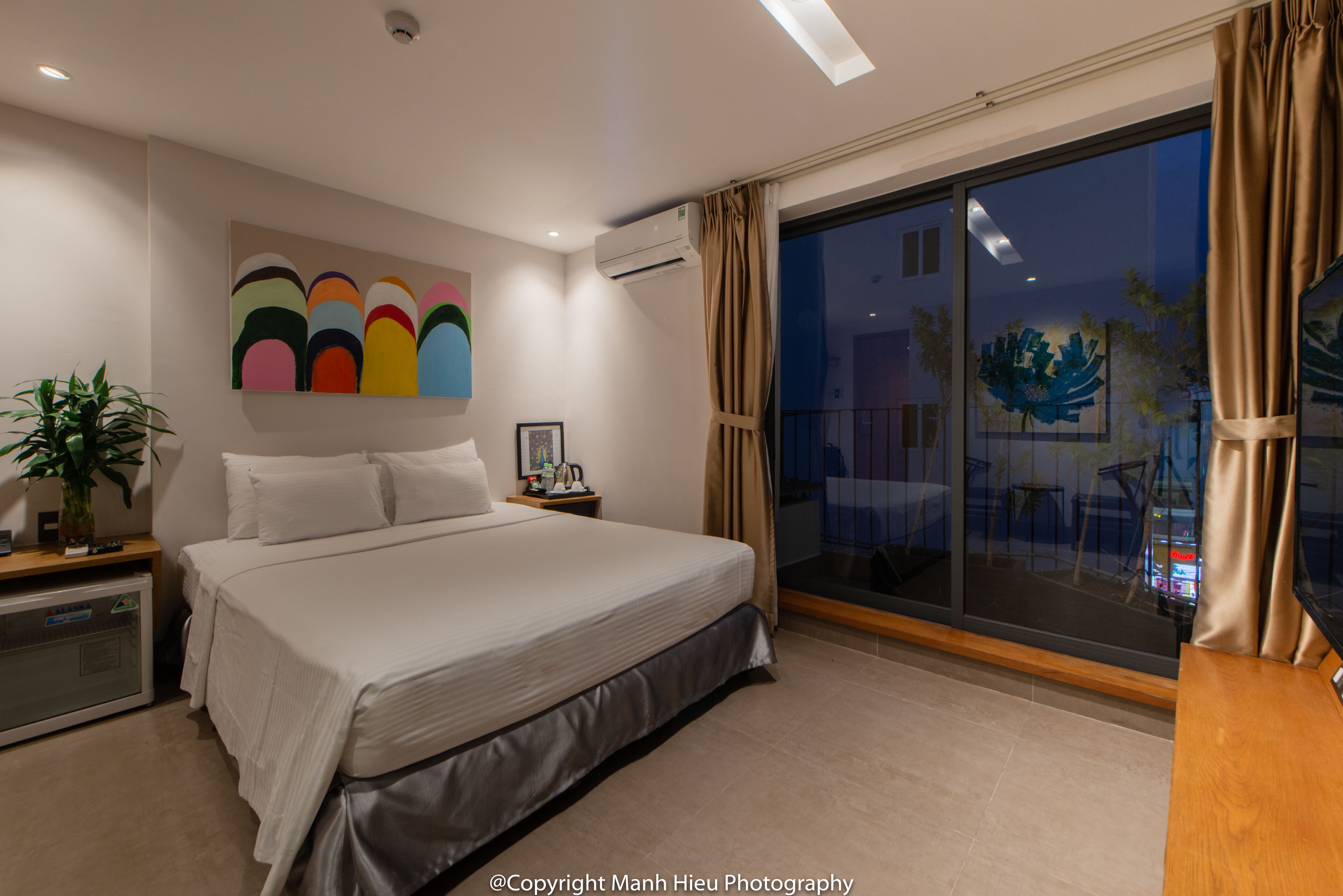 A modern hotel room with a large bed, a bedside table, a potted plant, and a view of the outside.
